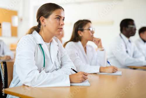 Girl medical student sitting at desk in classroom during lecture in medical college with group of students.