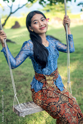 Canvas Print Balinese girl in traditional kebaya and batik sarong sitting on a swing in a tropical garden