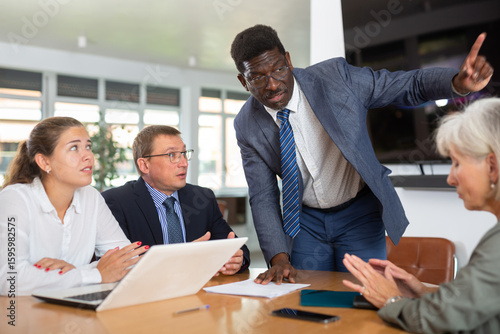 In conference room, African American man head of department speaks at meeting and reports unpleasant news to frowning colleagues. Secretary records leading moments of conversation in laptop
