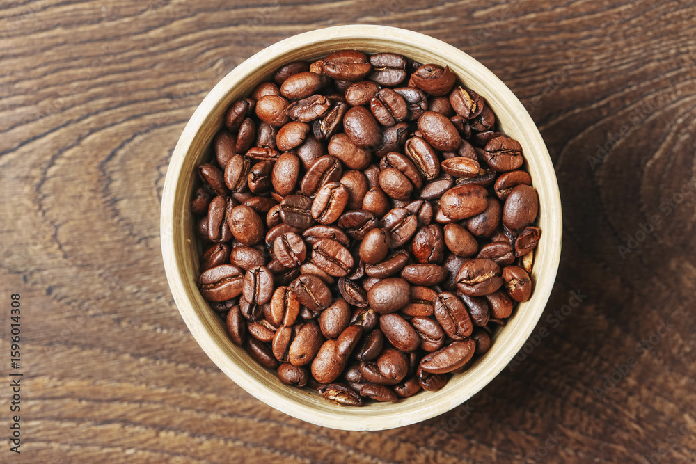 Fototapeta premium Top view of coffee beans. Wooden bowl on rustic table. Brown aromatic roasted grains. Freshly prepared natural caffeine. Morning energy concept. Simple food background with copy space.