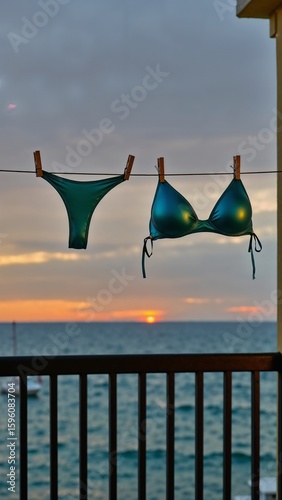 A shiny green bikini hangs on a clothesline on a balcony, drying against the backdrop of a beautiful, dramatic sunset over the ocean