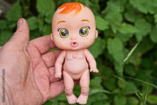 Fototapeta Naklejka Na Ścianę i Meble -  hand holds one small brown plastic toy doll on the street against the backdrop of green vegetation
