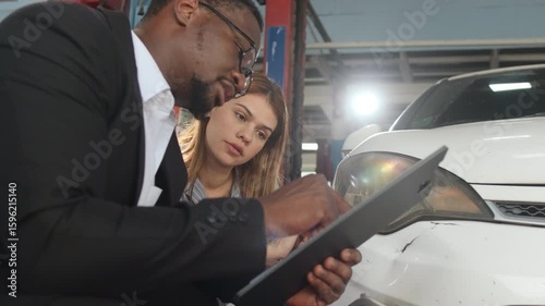 Warranty service business, Black male insurance agent inspector records crash car damage bumper, checks list and explains claim comprehensive of wreck fix to White female customer at auto garage.