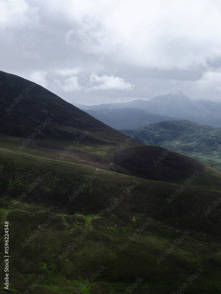 Fototapeta premium Mountains covered in green vegetation recede into the distance. The sky is grey with cloud cover over the scenic landscape of the Coomloughra Horseshoe in County Kerry.