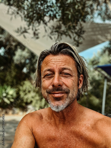 Smiling tanned man with stubble beard and blond hair enjoying summer garden with olive trees.
