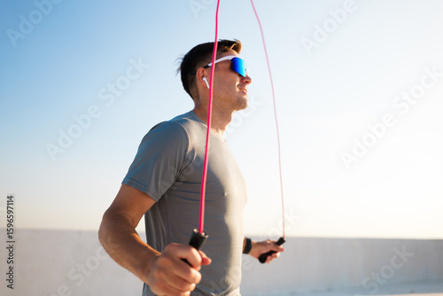 Fotografie Man skipping rope on rooftop at sunset wearing sunglasses and earphones