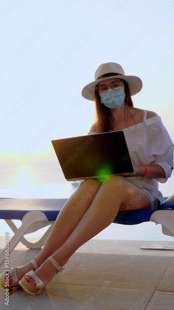 Young woman in protective mask, sun hat and summer clothes, working on laptop, sitting on sun lounger by the pool. work remotely. new normal. Life after Covid19.