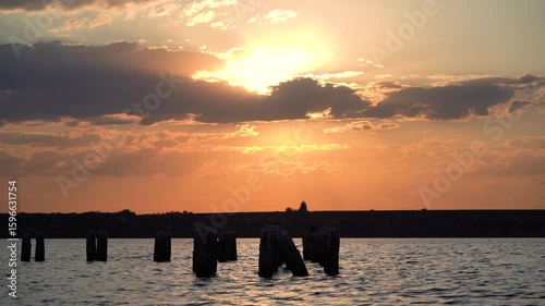 Evening sun dips behind the clouds as wooden pier remnants cast shadows on the water, evoking a sense of stillness and fading light 