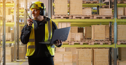 Valokuva Warehouse supervisor uses a laptop and two-way radio to manage inventory operations among stacked wooden crates on industrial shelving