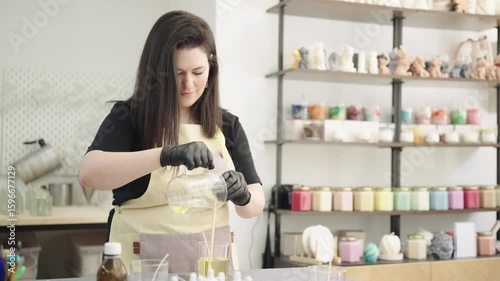 Woman carefully pouring melted wax into glass jars for candle making - small business concept