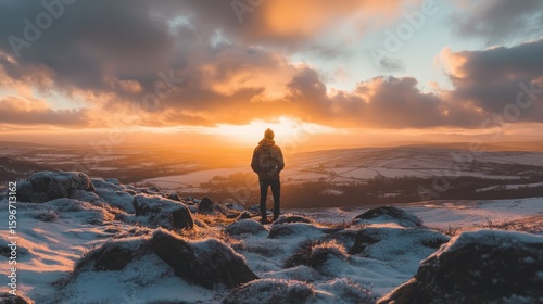 A solitary figure stands on rocky terrain, gazing at a vibrant sunset over a snowy landscape