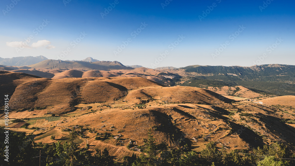 Fototapeta premium Rolling hills at the foot of Gran Sasso, Abruzzo, Italy, captured at sunset with warm golden light, soft shadows, and a serene landscape blending nature’s curves with mountain majesty.