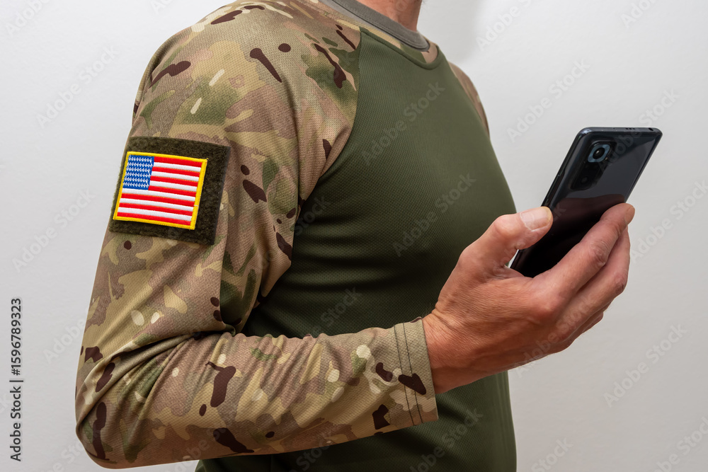 Fototapeta premium A US serviceman in camouflage with a US flag on his sleeve and a smartphone in his hand.