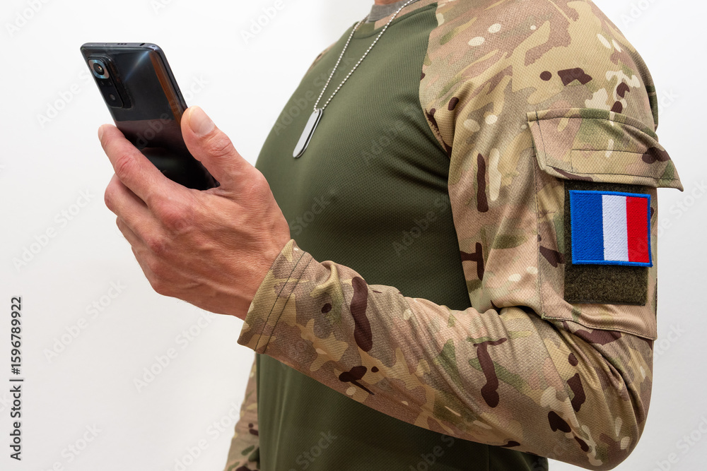 Fototapeta premium A soldier in tactical uniform with a French flag patch and a smartphone.