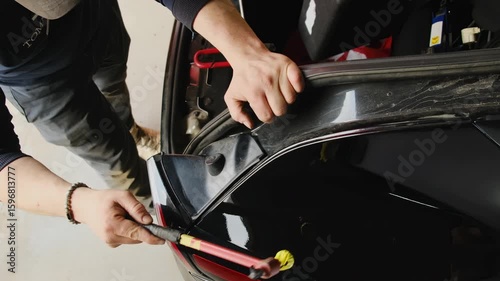 Man holds onto body of car hitting material with hammer. Master inspects surface of vehicle and monitors work results standing in hall
