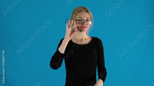 Enthusiastic model runs fingers near lips. Young woman asks to be silent as if closing mouth with key on blue background slow motion