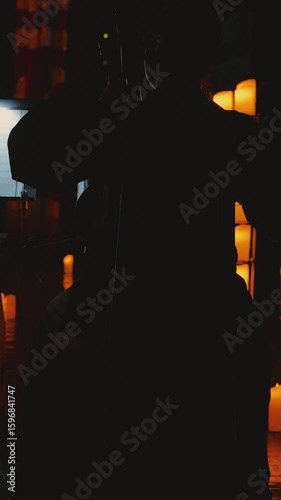 Close-up of the silhouette of a woman playing the cello in a dimly lit room surrounded by glowing candles. A candlelit concert