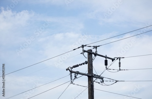 Wooden utility pole with high voltage power lines against blue sky with scattered clouds. Electrical wires crisscross, supported by insulators. Represents energy transmission, infrastructure,
