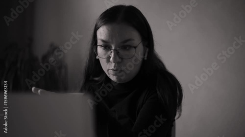 Black and white close-up of a thoughtful woman wearing glasses and earbuds, sitting indoors and looking at a screen. Emotional and introspective work or video call scene.