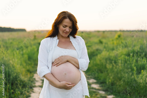 A pregnant woman stands in a green field. She gazes lovingly at her baby bump as the sun sets behind her, creating a serene atmosphere.