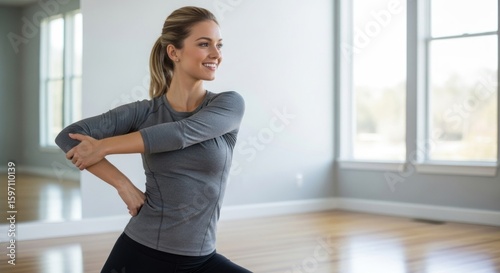 Free Download A woman in a gray workout top and black pants stretching in a spacious, well-lit room with large windows. - GFXLoad
