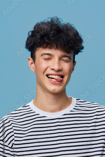 Smiling man with curly hair wearing a striped shirt playfully winking and sticking out his tongue against a solid blue background, showing fun and lively emotion.
