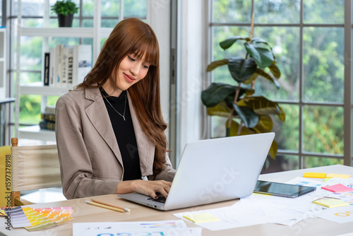 Young Asian woman working on laptop in creative modern workspace with color samples and documents on desk