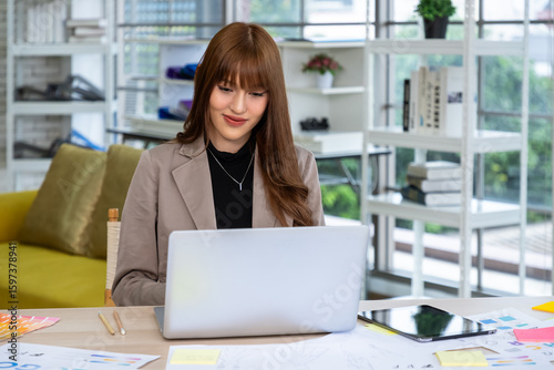 Young Asian designer woman working on laptop at modern creative desk with design sketches