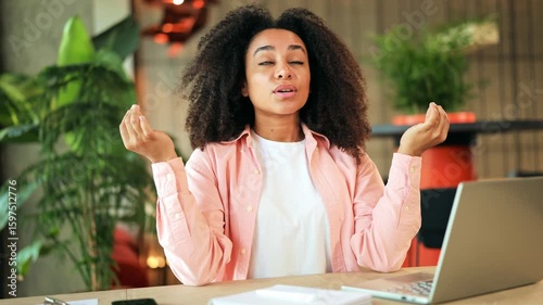 Peaceful woman with curly hair meditates calmly at her office desk, wearing a pink shirt and white t-shirt. She finds inner peace and relaxation, practicing mindfulness in a modern work environment.