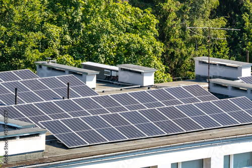Modern solar panels on a flat building roof with ventilation systems, surrounded by lush green trees. Clean energy and eco-friendly technology in an urban setting