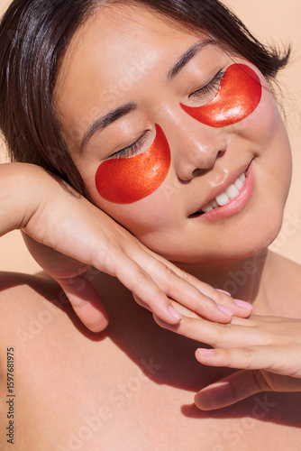 Asian woman with glowing skin, wearing red eye patches, is relaxing with a serene expression, showcasing a skincare routine in a bright, minimalistic setting