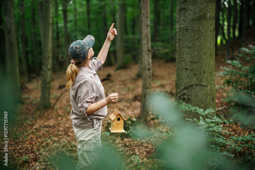 Woman ornithologist pointing toward spot for birdhouse installation in forest habitat, promoting bird nesting and protection