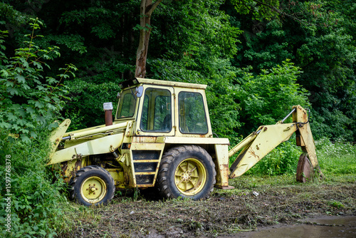 Old yellow backhoe loader parked at forest edge with overgrown vegetation