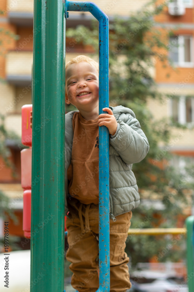 Obraz premium Vertical portrait of a small red-haired boy who looks over the fence of a playground in the city. Outdoor games