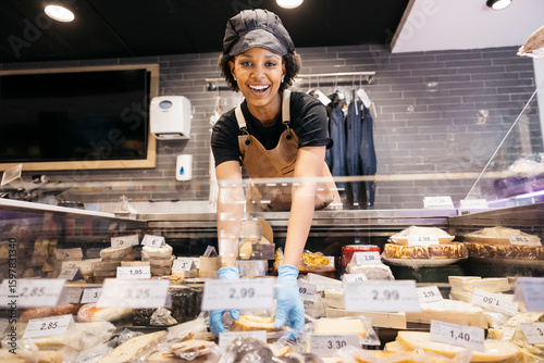 Bild auf Leinwand Young cheerful butcher wearing gloves arranging cheese in refrigerated display c