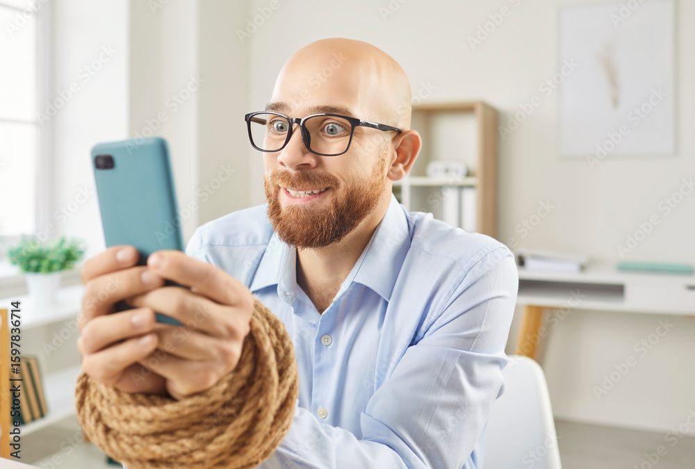 Portrait of a young obsessed business man employee sitting at the desk on workplace in office with his hands tied with rope addicted to mobile phone. Internet and smartphone addiction concept. Portrait of a young obsessed business man employee sitting at the desk on workplace in office with his hands tied with rope addicted to mobile phone. Internet and smartphone addiction concept.