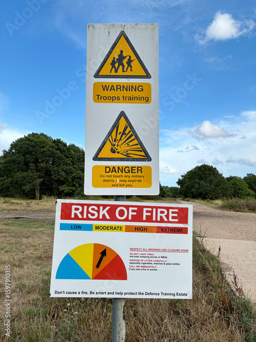 Warning signs reading 'Troops Training' and 'Risk of Fire' alert posted along woodland paths. Rural military training area natural setting with blue sky on summers day