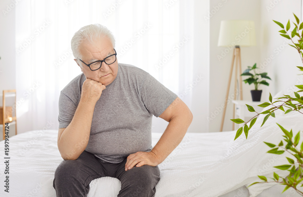 Sad senior man in grief sits in on bed in comfortable bedroom, deep in thought and thinking during the early morning of a new day. Emotions of despair, sadness and contemplation at home. Sad senior man in grief sits in on bed in comfortable bedroom, deep in thought and thinking during the early morning of a new day. Emotions of despair, sadness and contemplation at home.