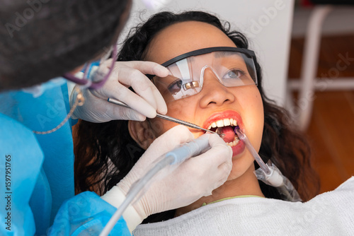 A young woman undergoes professional dental cleaning using an ultrasonic scaler, dental mirror, and suction during oral prophylaxis.