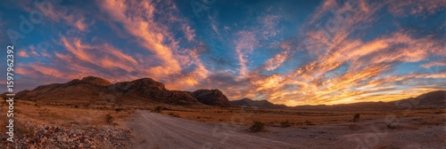 Dramatic desert landscape featuring a dirt road leading towards rocky mountains under a vibrant sunset sky filled with orange and blue clouds, capturing the beauty of nature.
