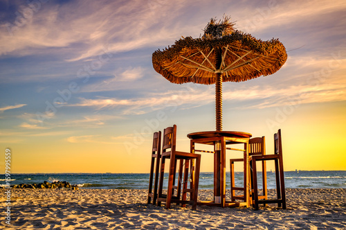 Billede på lærred Relaxing beach setup with thatched umbrella and wooden chairs under a clear blue