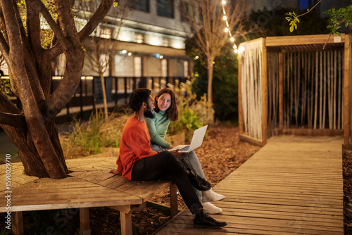 Happy multiracial male and female friends in casual clothes browsing assignment on netbook while sitting on bench in brightly illuminated park at night in park.