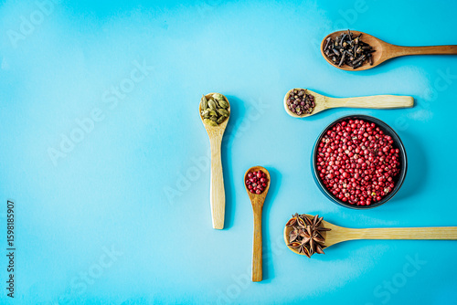 Fototapeta Naklejka Na Ścianę i Meble -  Wide flat-lay of wooden spoons filled with pink pepper, cardamom, cloves, star anise and Sichuan pepper framing a bowl of pink peppercorns on bright turquoise backdrop with ample copy space