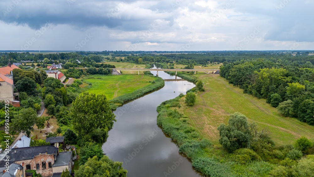 Fototapeta premium Aerial panorama of the Warta River in the town of Pyzdry. Green riverbanks, a bridge, and surrounding buildings create a peaceful rural landscape.