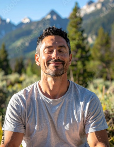 A man with closed eyes and a slight smile enjoys a peaceful moment outdoors in nature with mountains and trees in the background.