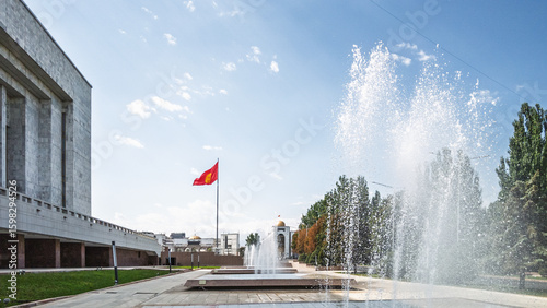 A panoramic view of Ala-Too Square in Bishkek, Kyrgyzstan, featuring the Kyrgyz flag, a modern government building, and a series of elegant fountains.  The image evokes a sense of civic pride and