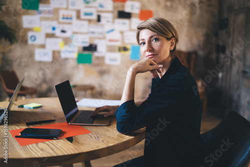 Smart female freelancer gazing at viewer while typing on laptop, showing clarity, confidence and digital independence in bright, tech-friendly creative office space