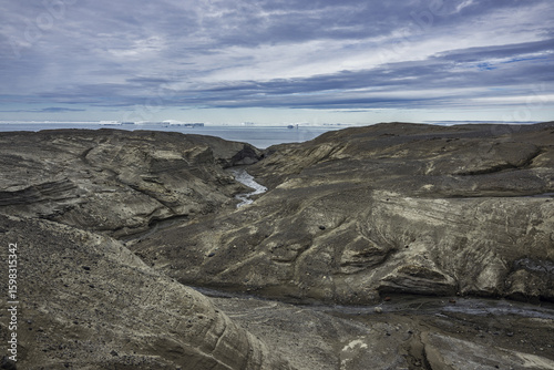 View of a rugged, desolate landscape with a stream cutting through dark rock formations under a sky streaked with clouds, Seymour Island, Antarctica.