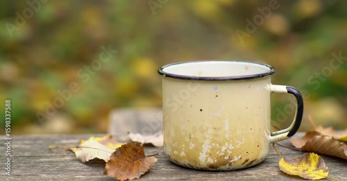 A vintage enamel mug sits on a rustic wooden table surrounded by autumn leaves, evoking a cozy fall atmosphere.