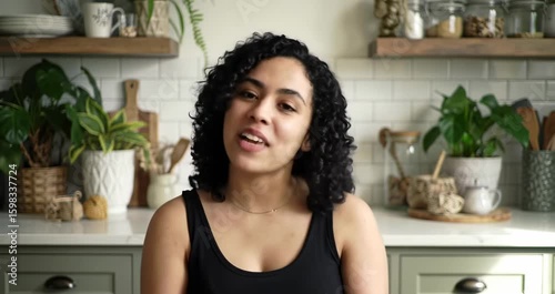 A joyful young woman with curly hair smiles warmly in a cozy kitchen setting filled with plants.